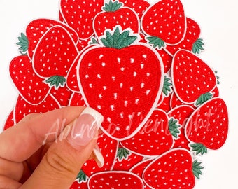 May include: A pile of red embroidered strawberry patches with white outlines and green leaves. The patches are arranged in a circle with one patch being lifted up by a hand.