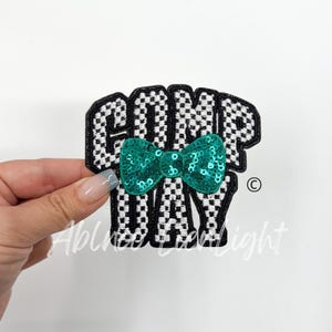May include: A black and white checkered fabric patch with the words "GOMP DAY" in white embroidery. A teal sequin bow is centered on the patch.