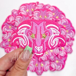 May include: Pink and white embroidered iron-on patch of an Aries zodiac sign with gold glitter stars. The patch is held in a hand with green nail polish.