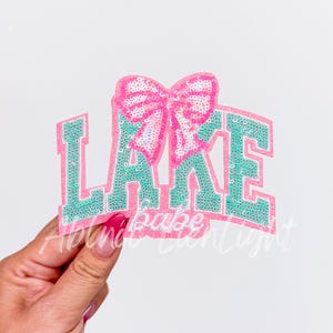 May include: A pink and green iron-on patch with the word "LAKE" in large, sparkly letters and a pink bow with white sequins in the center.