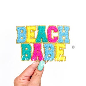 May include: A colorful felt applique with the words "Beach Babe" in gold glitter letters. The letters are in shades of blue, yellow, pink, and turquoise.