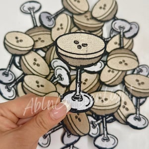 May include: A pile of embroidered iron-on patches featuring a martini glass with coffee beans. The patches are beige and brown with black outlines.
