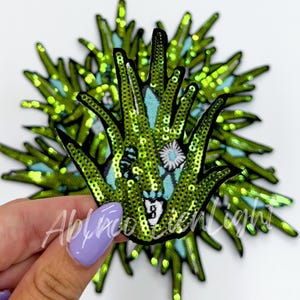 May include: Green sequined iron-on patch with a floral design. The patch is shaped like a hand with five fingers.
