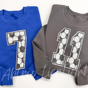 May include: Two sweatshirts with soccer-themed numbers. One is royal blue with the number 7 and the other is gray with the number 11.