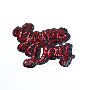 May include: A sequined patch with the words "Game Day" in red script. The letters are filled with red sequins and outlined with a black glitter border. The patch is on a white background.