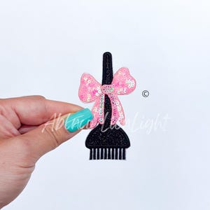 May include: Pink sequined bow on a black hair brush. The brush has a handle and a row of bristles.