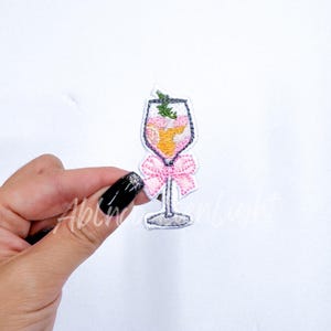 May include: Embroidered iron-on patch of a pink cocktail glass with a pink bow. The glass is filled with a light orange liquid and has a green sprig on top.