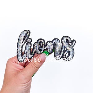 May include: A black and silver sequin iron-on patch that says "lions" in a cursive font.