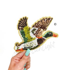 May include: A green, brown, and white sequined iron-on patch of a flying duck. The duck has a gold outline.