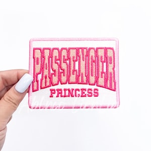 May include: A rectangular, embroidered patch with the words "PASSENGER PRINCESS" in pink and light pink lettering. The patch has a white background and a pink border. The patch is held by a hand with a white nail polish.