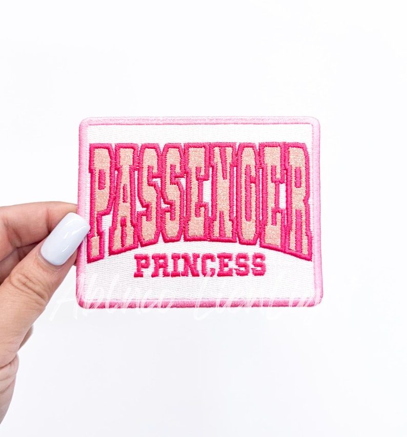Puede incluir: Parche rectangular bordado con las palabras "PASSENGER PRINCESS" en rosa y rosa claro. El parche tiene un fondo blanco y un borde rosa. El parche est&aacute; sujeto por una mano con esmalte de u&ntilde;as blanco.