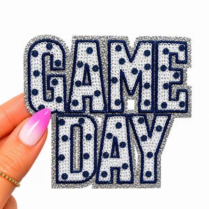 May include: A decorative iron-on patch featuring the words GAME DAY in bold, block letters. The letters are covered in white sequins with dark blue polka dots and are outlined with a thick, shimmering silver border for a textured appearance.
