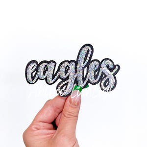 May include: A black iron-on patch with the word "eagles" in silver glitter sequins.