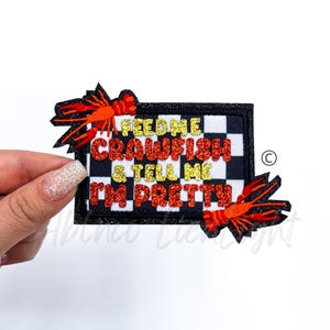 May include: A black and white checkered patch with the text "Feed me crawfish & tell me I'm pretty" in red and gold glitter. Two red crawfish are on either side of the patch.