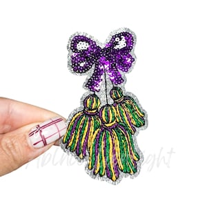 May include: A sequined purple bow with three tassels in green, yellow, and purple. The design has a silver glitter border. The bow and tassels are connected by thin silver strings. The design is on a white background.