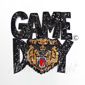 May include: Black sequined "GAME DAY" patch featuring a roaring bear with gold and black sequins. The bear has a red mouth and white teeth. The patch is on a white background.
