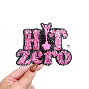 May include: A pink sequined patch with the words "HIT ZERO" in a stylized font. The patch has a black glitter border and a pink figure. The design is on a white background.