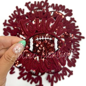 May include: Maroon football-shaped applique with the word "Touchdown" in cursive script. The football is embellished with sequins and white stitching. A hand holds the applique against a white background.