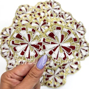 May include: A close-up of a pile of red, white, and gold glitter cheer pom poms. The pom poms are embroidered with a white and gold swirl design.
