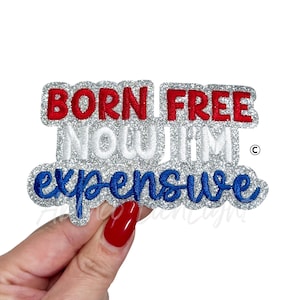 May include: A glittery patch with the text "BORN FREE NOW I'M expensive" in red, white, and blue. The patch is held by a hand with red nail polish.