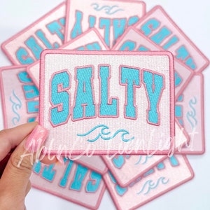 May include: A stack of pink and white embroidered patches with the word "SALTY" in blue lettering and a wave design.