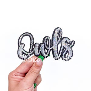 May include: A black sequined patch with the word "Quofs" in a cursive font. The letters are outlined in black and filled with silver sequins. The patch is held against a white background.