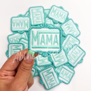 May include: A pile of teal blue iron-on patches with the word "MAMA" embroidered in white. The patches are in a square shape with a decorative border.