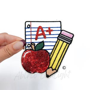 May include: A red apple with a green stem and leaves, a yellow pencil with a pink eraser, and a white sheet of paper with blue lines and the text "A+" in red glitter.