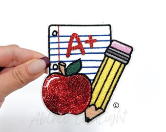 May include: A red apple with a green stem and leaves, a yellow pencil with a pink eraser, and a white sheet of paper with blue lines and the text "A+" in red glitter.