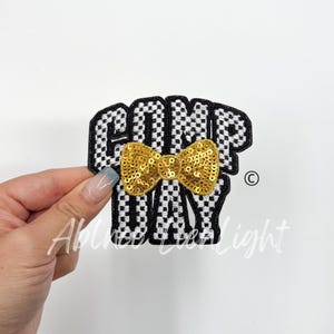 May include: Black and white checkered iron-on patch with the text "COMP DAY" and a gold sequin bow.