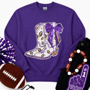 May include: A purple crewneck sweatshirt featuring a large central graphic of white cowboy boots decorated with brown footballs, gold stars, and a prominent purple sequined bow. The design includes the word TOUCHDOWN written on the boot.