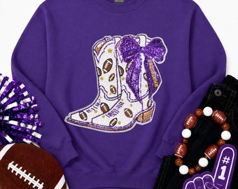 May include: A purple crewneck sweatshirt featuring a large central graphic of white cowboy boots decorated with brown footballs, gold stars, and a prominent purple sequined bow. The design includes the word TOUCHDOWN written on the boot.