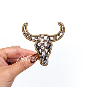 May include: A brown and white buffalo skull patch with a plaid pattern. The patch has a gold glitter outline and is held by a hand. The text "Ablaze Moon Light" is written on the patch.
