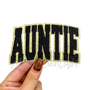 May include: A black and gold glitter patch with the word "AUNTIE" in a bold, collegiate-style font. The patch has a black embroidered design with gold glitter accents, perfect for adding to clothing or accessories.