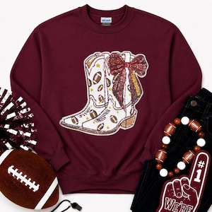May include: A maroon crewneck sweatshirt featuring a large graphic of two white cowboy boots decorated with small brown footballs and gold stars. A large, glittery red bow is attached to the boots, with the word TOUCHDOWN printed in gold.