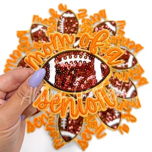 May include: A stack of iron-on patches featuring a football design and the text "Mom of a Senior". The football is decorated with brown and pink sequins.