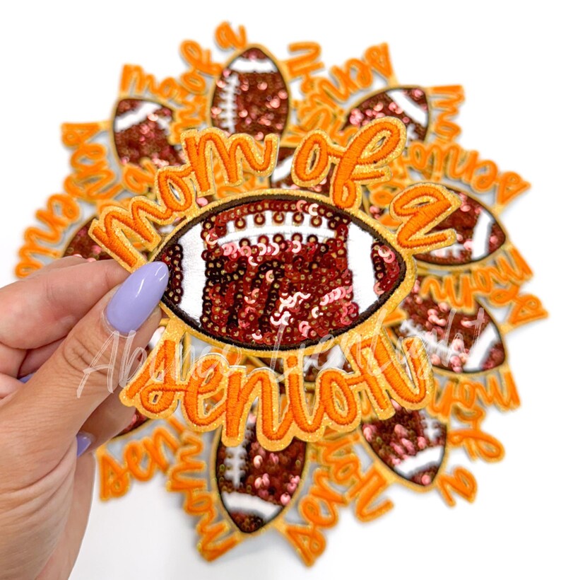 May include: A stack of iron-on patches featuring a football design and the text "Mom of a Senior". The football is decorated with brown and pink sequins.
