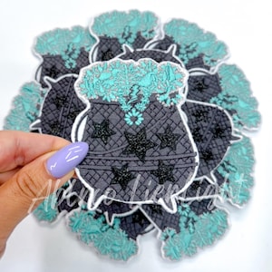 May include: A stack of black and teal cauldron iron-on patches with a silver glitter design. The cauldron is filled with teal glitter and has a black lightning bolt in the center. The cauldron is surrounded by black stars.