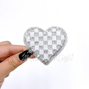 May include: A silver heart-shaped patch with a checkered pattern and sequin trim. The patch is held against a white background. The patch has a glittery appearance.