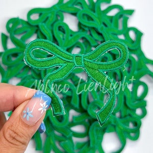 May include: A pile of green felt fabric bow appliques. The bows are about 2 inches wide and have a satin-like finish.