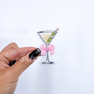 May include: A white embroidered patch of a martini glass with a green olive and a pink bow.