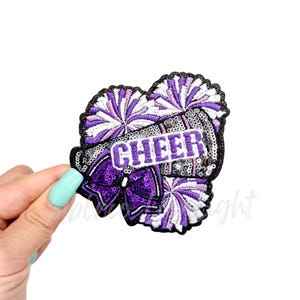 May include: Purple and white cheerleading pom poms with a silver sequined megaphone with the word "CHEER" on it.