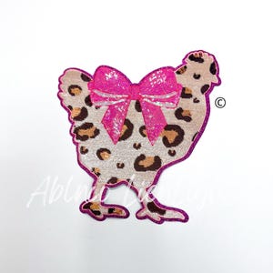 May include: A pink and brown sequined chicken applique with a pink bow. The chicken is standing on two legs and has a brown and white leopard print pattern.