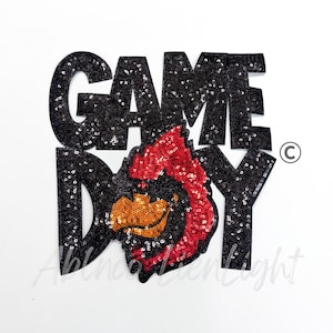 May include: Black sequined "GAME DAY" applique with a red and orange cardinal bird design. The applique is on a white background. The cardinal bird is in the center of the applique.