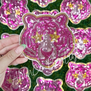 May include: Pink and gold sequined iron-on patches of tiger faces with gold stars for eyes.
