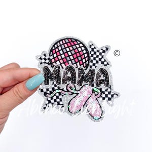 May include: A glittery patch with the word "MAMA" in black sequins. The patch features a pink and black checkered disco ball, pink ballet shoes, and a silver glitter border with a black and white checkered pattern.