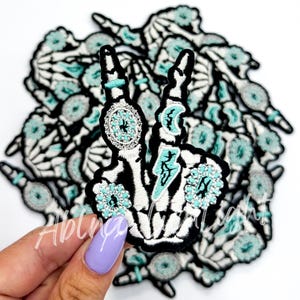 May include: A pile of embroidered iron-on patches featuring a skeleton hand making the peace sign. The hand is decorated with turquoise and silver glitter.