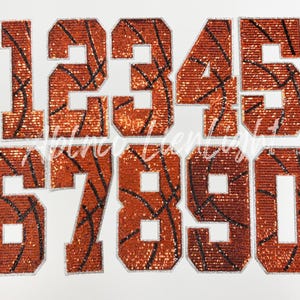 May include: A set of iron-on numbers, 0-9, with a basketball design. The numbers are orange with black lines, resembling a basketball, and outlined in silver. The numbers are displayed on a white background.