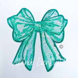 May include: A large, teal sequined bow with long, flowing tails. The bow is embellished with iridescent sequins, creating a shimmering effect. The text "Abinco Lienlight" is printed on the lower left side of the bow.