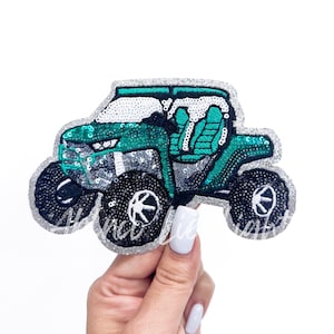 May include: A sequined patch in the shape of a teal and silver off-road vehicle. The patch has black wheels with white accents and a silver glitter border. The vehicle's interior is teal, and the roof is white.